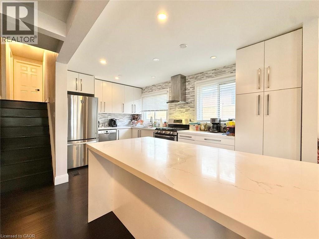 Modern kitchen featuring a large center island with a light-toned countertop, sleek white cabinetry, stainless steel appliances, and a textured tile backsplash - 3135 Palmer Drive, Burlington, ON