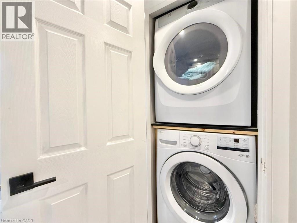 Integrated laundry station featuring a white stackable washer and dryer, a wood-finish platform, and a paneled door with a matte black lever handle - 3135 Palmer Drive, Burlington, ON