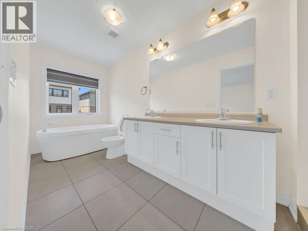 Spacious bathroom featuring a freestanding soaking tub, dual vanity with undermount sinks, stone-finish countertop, large wall-mounted mirror, and neutral tile flooring - 76 Grassbourne Avenue, Kitchener, ON