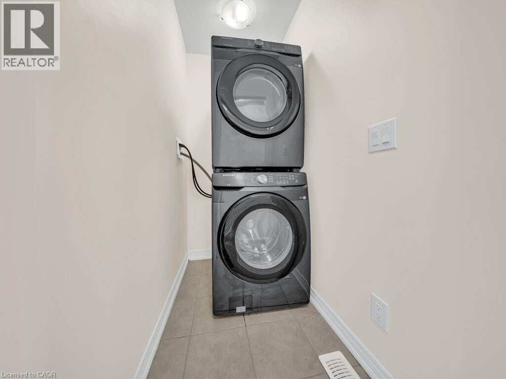 Dedicated laundry area featuring a stacked washer and dryer unit, neutral wall paint, white trim, and tiled flooring - 76 Grassbourne Avenue, Kitchener, ON