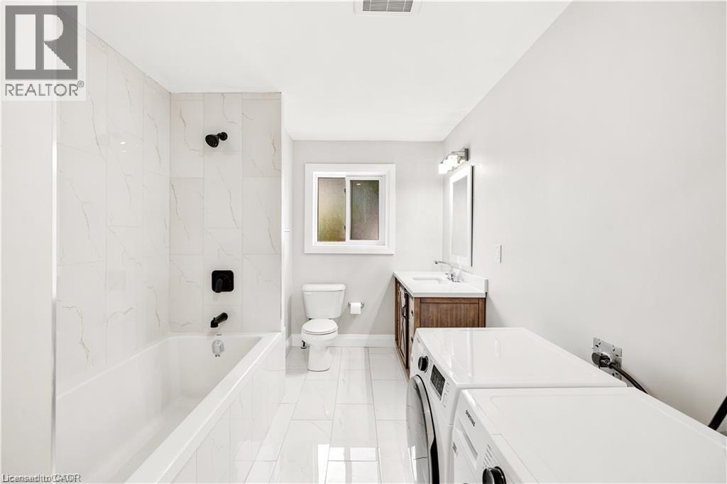 Modern bathroom featuring a white bathtub with matte black fixtures, white large format tile, a wood-finish vanity with a white countertop, and an integrated washer and dryer - 129 St George Street, Kitchener, ON