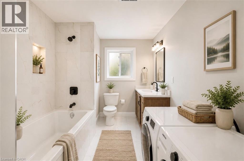 Bathroom featuring a built-in tub with matte black fixtures and a tiled surround, a window providing natural light, a vanity with wood-finish cabinetry and a single-basin sink, and a stacked washer an - 129 St George Street, Kitchener, ON