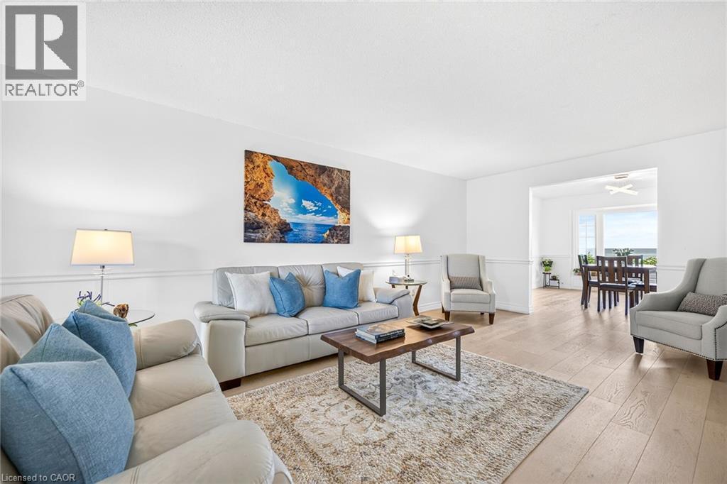 Bright living area featuring light wood-finish flooring, white walls with wainscoting, and an open doorway to a dining area with expansive windows - 15 Cheval Drive, Grimsby, ON - Indoor Photo Showing Living Room