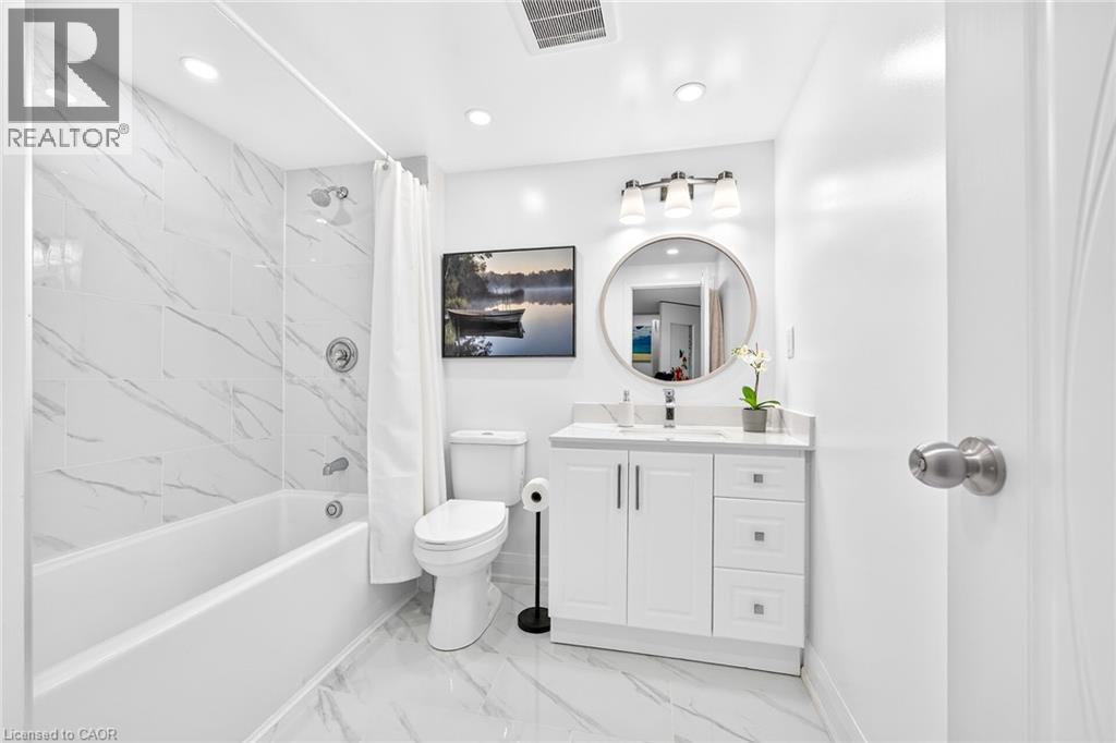 Bathroom featuring white marble-look wall and floor tile, a white vanity with a light-colored countertop, and a circular mirror with an overhead three-light fixture - 15 Cheval Drive, Grimsby, ON - Indoor Photo Showing Bathroom