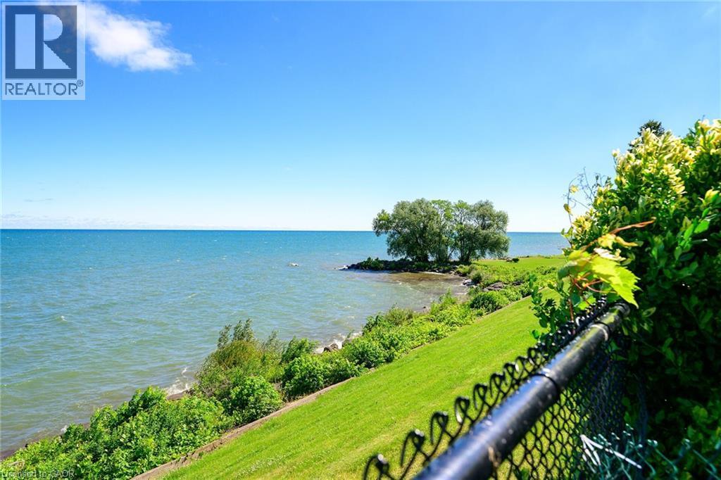 Expansive waterfront setting featuring a grassy slope, mature trees, and a black chain-link fence - 15 Cheval Drive, Grimsby, ON - Outdoor With Body Of Water With View