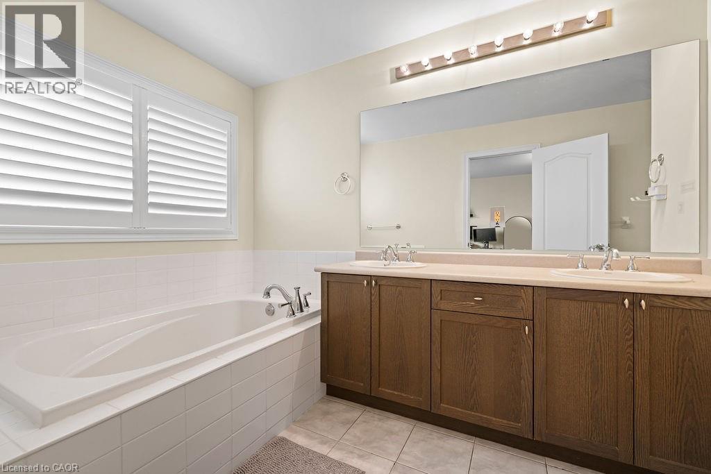 Built-in soaking tub with white tile surround, accompanied by a double vanity featuring wood-finish cabinetry, integrated sinks, a light-toned countertop, and an expansive wall-mounted mirror - 4670 Thomas Alton Boulevard, Burlington, ON