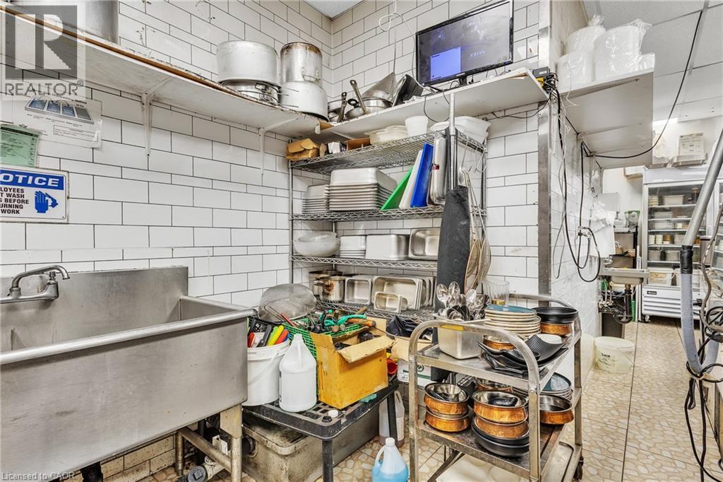 Commercial-grade stainless steel sink with wall-mounted faucet, white subway tile walls, open metal shelving, and commercial refrigerator visible in the background - 155 Highland Road E, Kitchener, ON