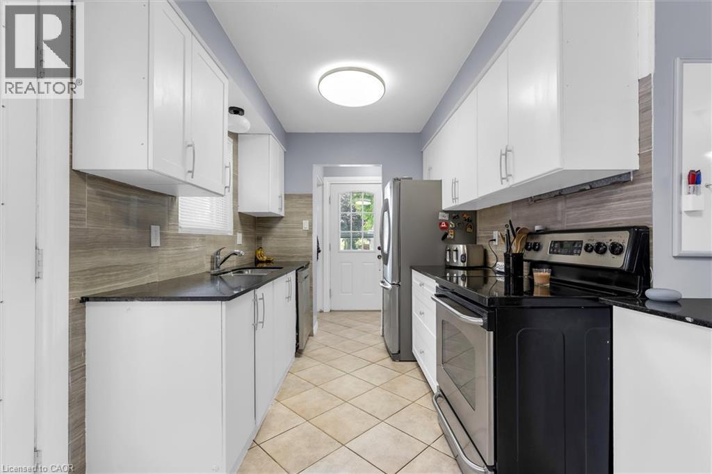 Galley kitchen featuring abundant white cabinetry, dark countertops, a tile backsplash, stainless steel appliances, and tile flooring - 338 East 24Th Street, Hamilton, ON
