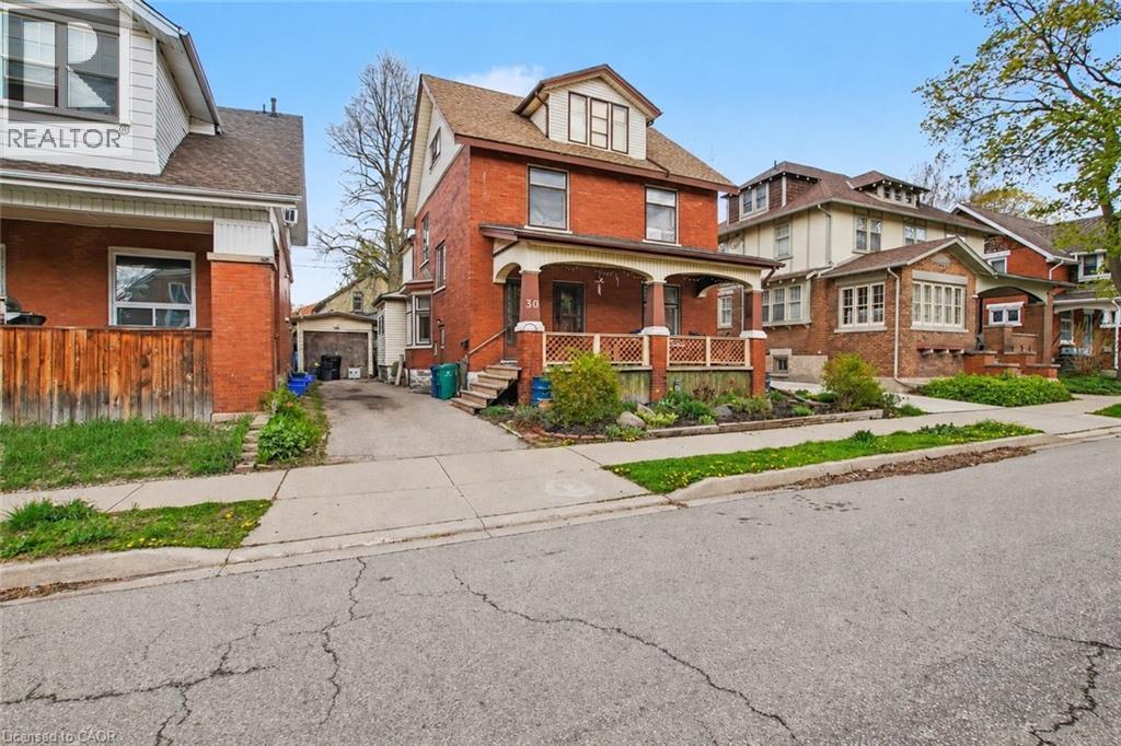 Brick exterior with a covered front porch featuring decorative railing, a paved driveway, and mature landscaping - 30 Hohner Avenue, Kitchener, ON - Outdoor With Facade