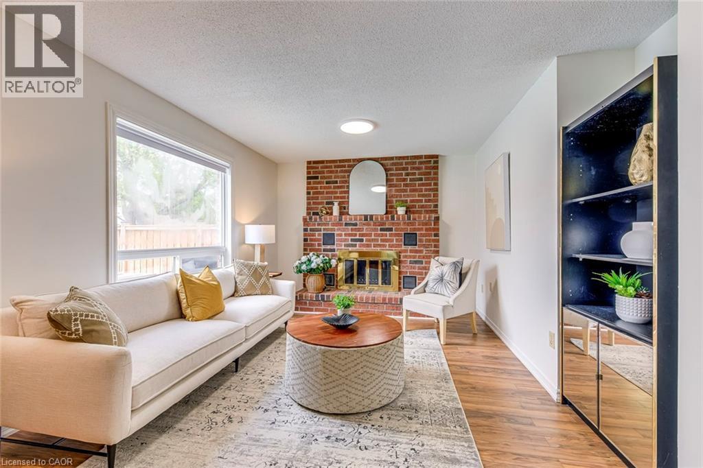 Living area featuring a brick fireplace with a brass firebox, wood-finish flooring, a large picture window, and a built-in shelving unit with mirrored doors - 43 Niska Drive, Waterdown, ON - Indoor Photo Showing Living Room With Fireplace
