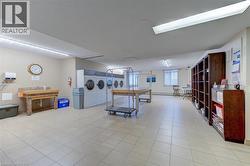 Spacious common area featuring an array of large-capacity washing machines, light-toned tile flooring, built-in shelving, a utility sink, and ample overhead lighting - 