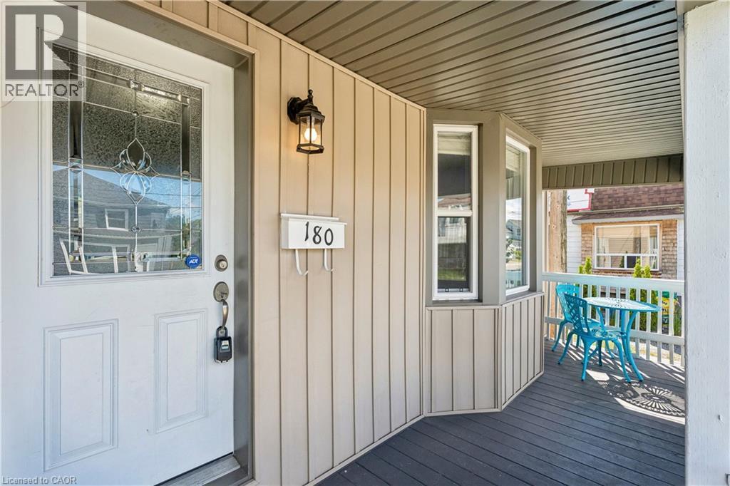 Covered front porch featuring a white entry door with decorative glass inlay, an exterior wall lantern, bay windows, and a dark-toned composite deck - 180 Campbell Avenue, Hamilton, ON - Outdoor With Deck Patio Veranda With Exterior
