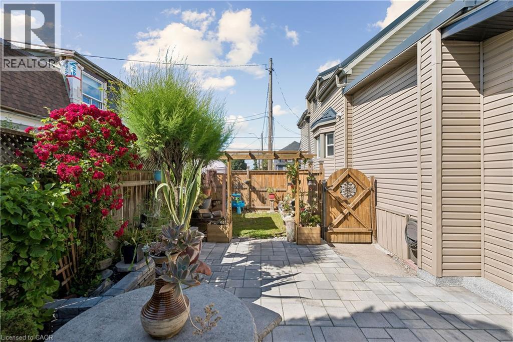 Paver patio with a decorative wooden gate and vibrant red flowering shrubs - 180 Campbell Avenue, Hamilton, ON - Outdoor With Deck Patio Veranda