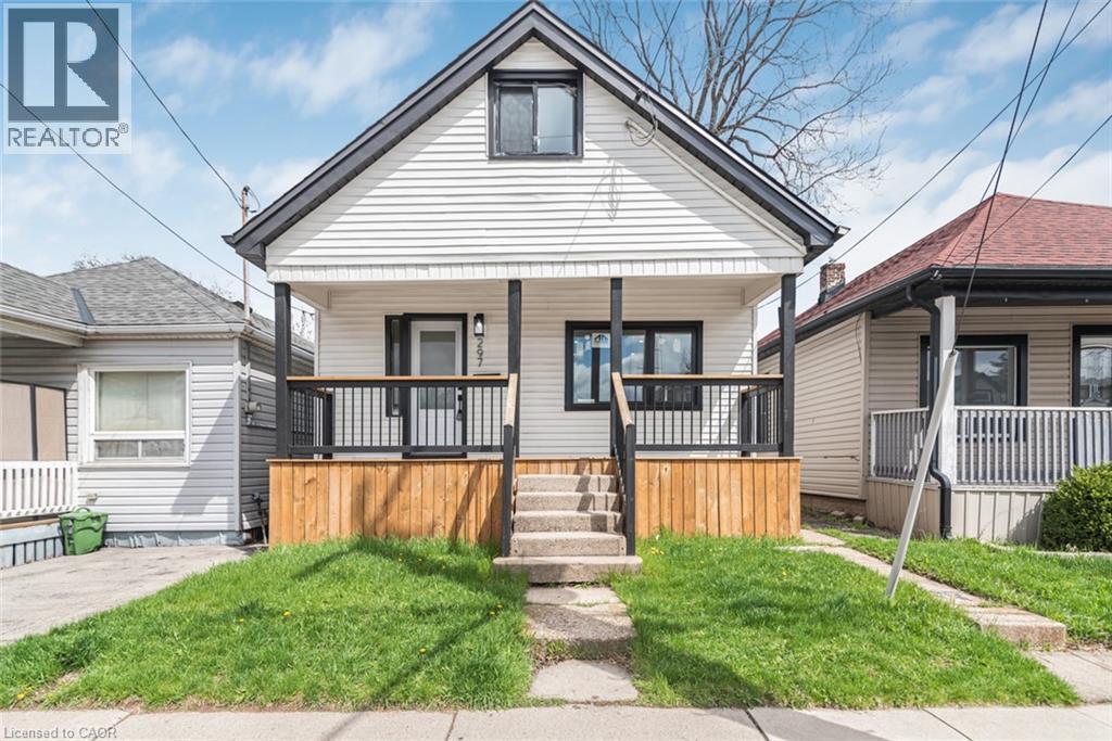 Gable-roof property featuring white horizontal siding, black trim accents, a covered front porch with a wood-paneled base, black railings, and concrete steps - 297 Britannia Avenue, Hamilton, ON