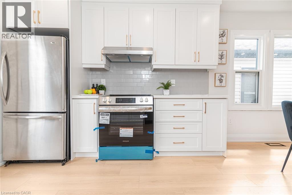 Contemporary kitchen featuring stainless steel appliances, white cabinetry with gold-tone hardware, a subway tile backsplash, light-tone wood-finish flooring, and natural light from multiple windows - 297 Britannia Avenue, Hamilton, ON