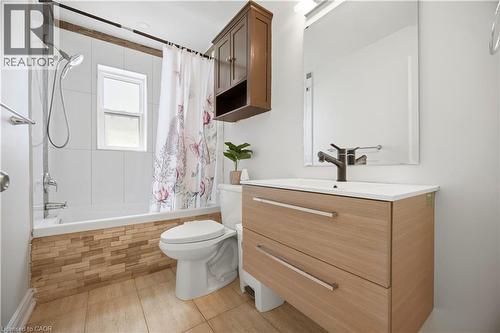 Bathroom featuring a modern vanity with integrated sink, a rectangular mirror with integrated lighting, a tub with white tile surround, and wood-finish flooring - 71 Beland Avenue N, Hamilton, ON 
