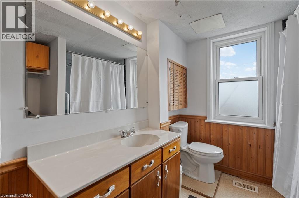 Bathroom featuring a single vanity with a white countertop and wood-finish cabinetry, a large wall-mounted mirror, a window with natural light, wood-paneled wainscoting, and a built-in louvered cabine - 2 Mill Street E, York, ON