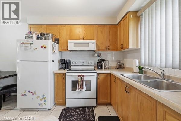 Kitchen featuring wood-finish cabinetry, white appliances, a double basin stainless steel sink, tile backsplash, and light-toned countertops - 16 Harrison Avenue, Hamilton, ON