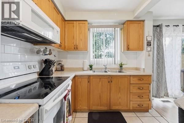 Kitchen featuring wood-finish cabinetry, a white subway tile backsplash, a double basin sink with window, white appliances, and light-toned floor tiling - 16 Harrison Avenue, Hamilton, ON