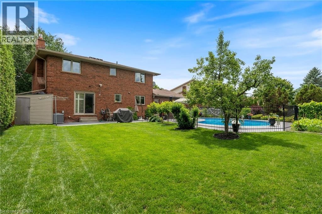 Expansive backyard featuring a manicured lawn, an in-ground swimming pool with a black iron fence, mature trees, a red brick exterior, and a patio area - 7 Thomson Road, Simcoe, ON