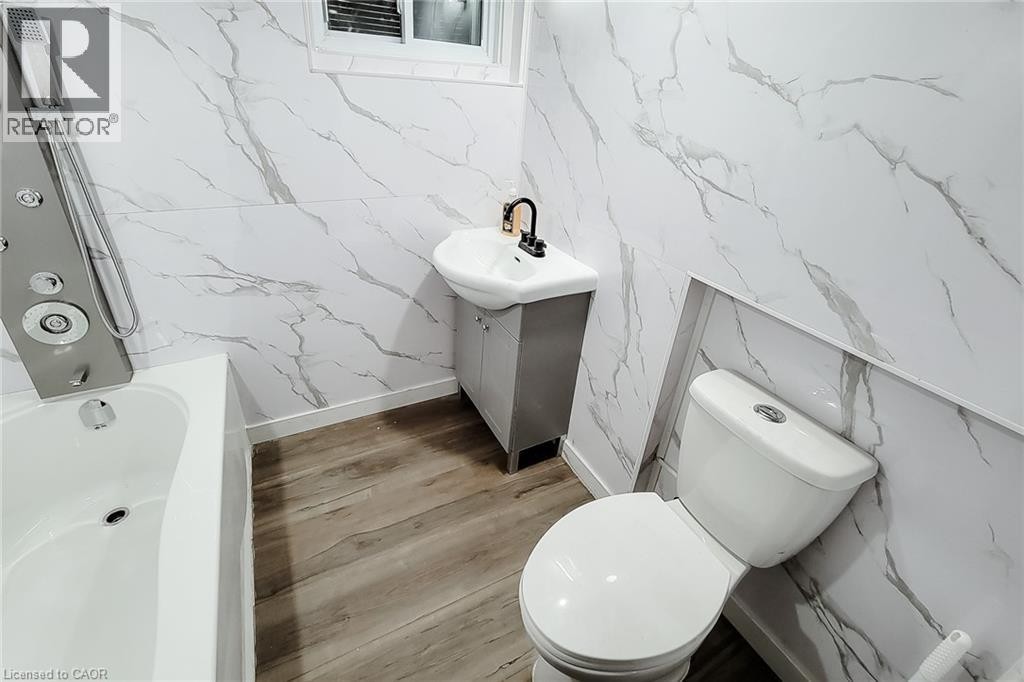 Bathroom featuring white marbled wall panels, wood-finish flooring, white soaking tub with a multi-function shower panel, and a white toilet - 78 Hillyard Street, Hamilton, ON