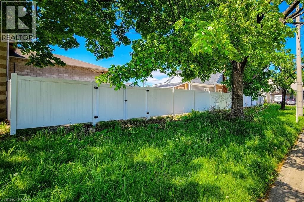 White privacy fencing borders a grassy expanse, complemented by mature trees and a paved sidewalk - 78 Hillyard Street, Hamilton, ON