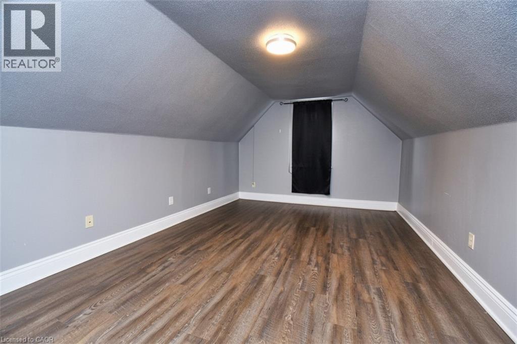 Spacious room featuring wood-finish flooring, light grey walls, white baseboards, and a sloped ceiling with a flush-mount light fixture - 242 James Street S, Hamilton, ON