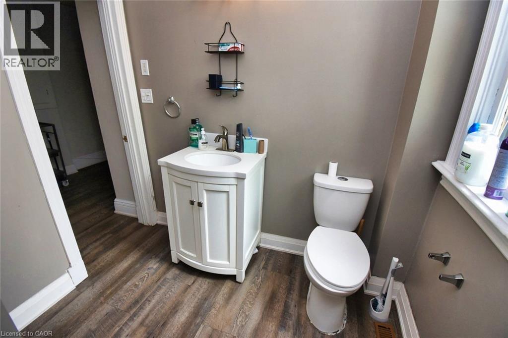 Bathroom featuring wood-finish flooring, a white vanity with an integrated sink, a white toilet, and a window with white trim - 242 James Street S, Hamilton, ON
