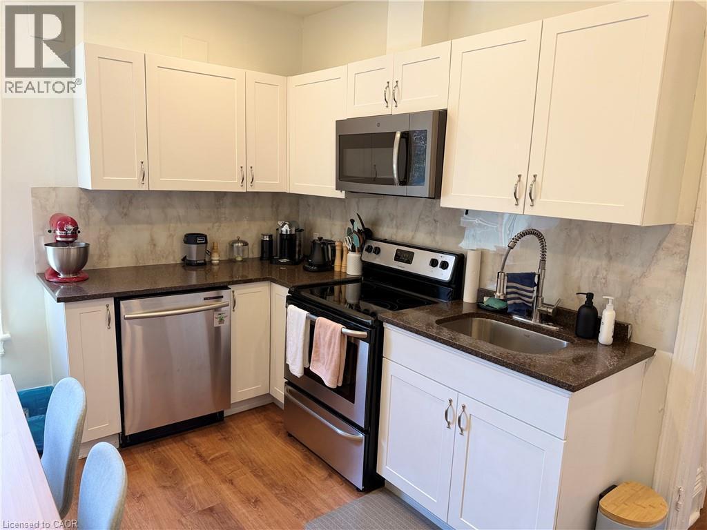 Corner kitchen featuring white upper and lower cabinetry, dark countertops, a stainless steel dishwasher, a stainless steel range oven, and wood-finish flooring - 207 Charlton Avenue W, Hamilton, ON