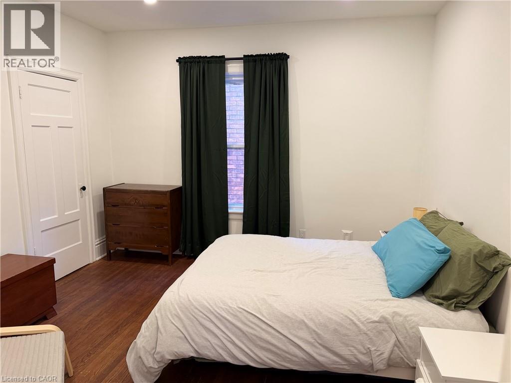 Room featuring wood-finish flooring, a window with dark curtain panels, and a white paneled door - 207 Charlton Avenue W, Hamilton, ON