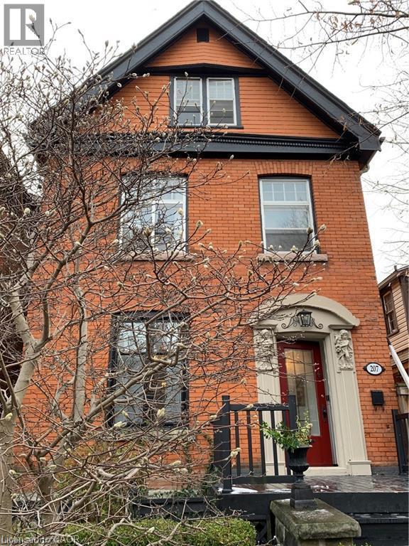 Charming brick Colonial featuring a gabled roofline with dark architectural detailing, multi-pane double-hung windows, and a covered entry portico with red door and black iron railings - 207 Charlton Avenue W, Hamilton, ON