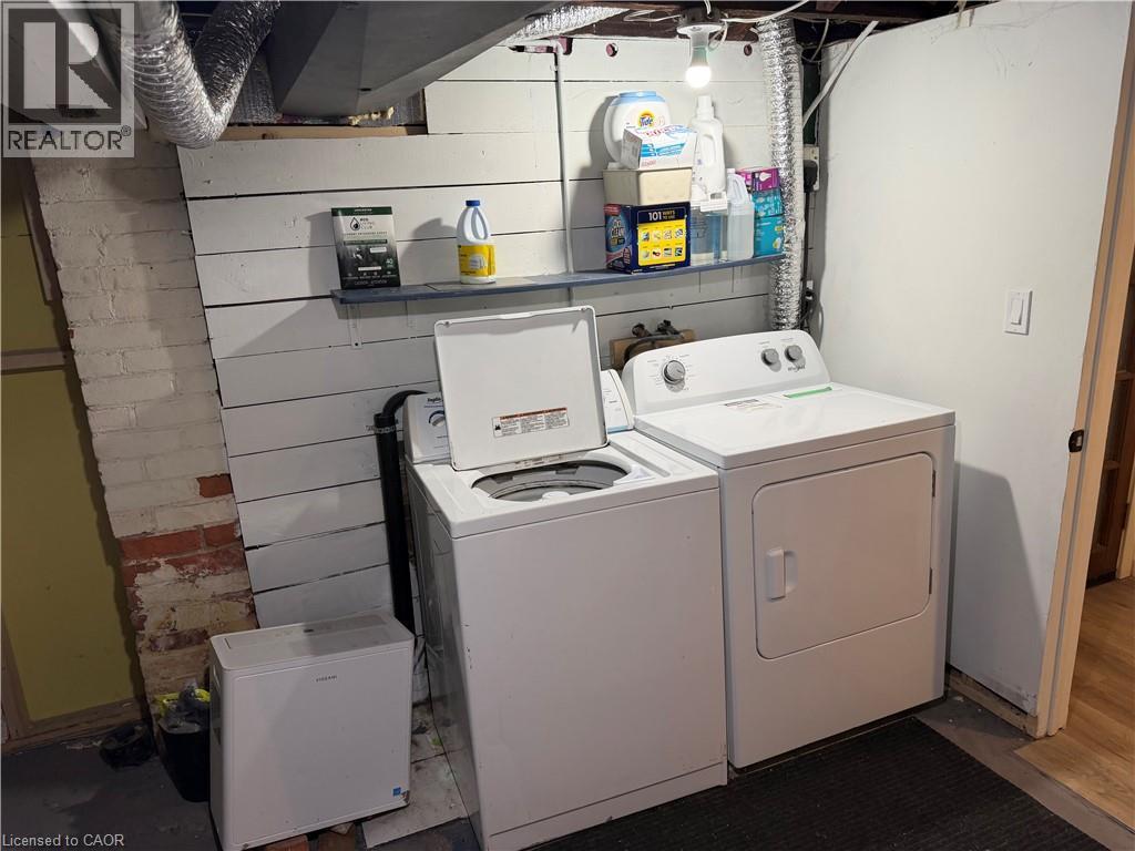 Dedicated laundry area featuring a white washing machine and dryer, exposed brick wall, and overhead shelving - 207 Charlton Avenue W, Hamilton, ON