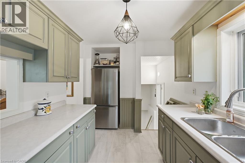 Galley kitchen featuring light green cabinetry, light countertops, wood-finish flooring, a stainless steel refrigerator, and a double basin sink - 81 Vale Avenue, St. Catharines, ON