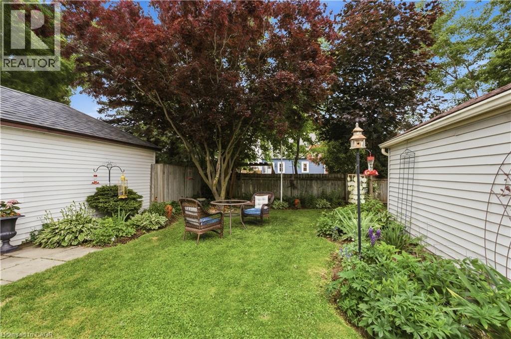 Lush backyard featuring a mature red-leafed tree, manicured lawn, garden beds, white-paneled exterior structures, and a wooden privacy fence - 81 Vale Avenue, St. Catharines, ON