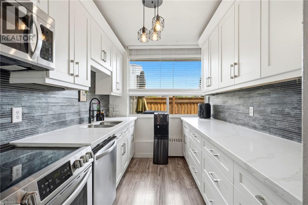 Galley kitchen featuring white shaker cabinetry, stainless steel appliances, a tile backsplash, light-toned countertops, and wood-finish flooring - 15 Nicklaus Drive Unit# 17, Hamilton, ON