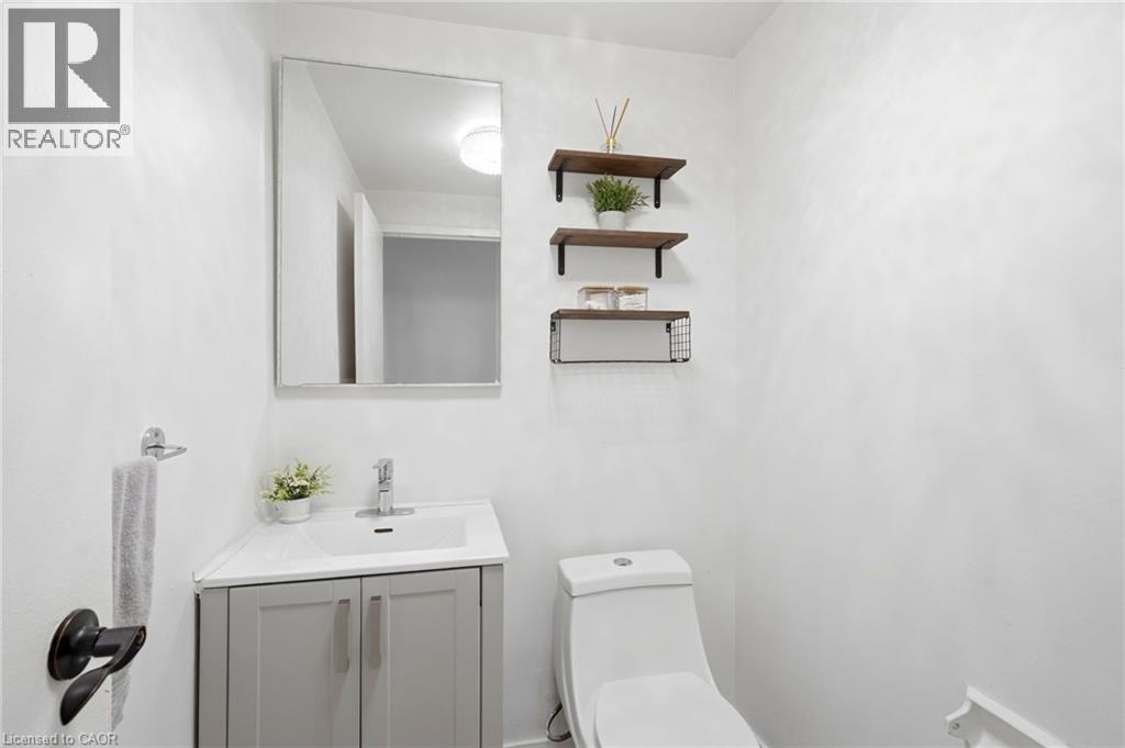 Compact bathroom featuring a corner vanity with a white countertop and gray cabinetry, a wall-mounted mirror, a two-piece toilet, and three wall-mounted shelves - 15 Nicklaus Drive Unit# 17, Hamilton, ON