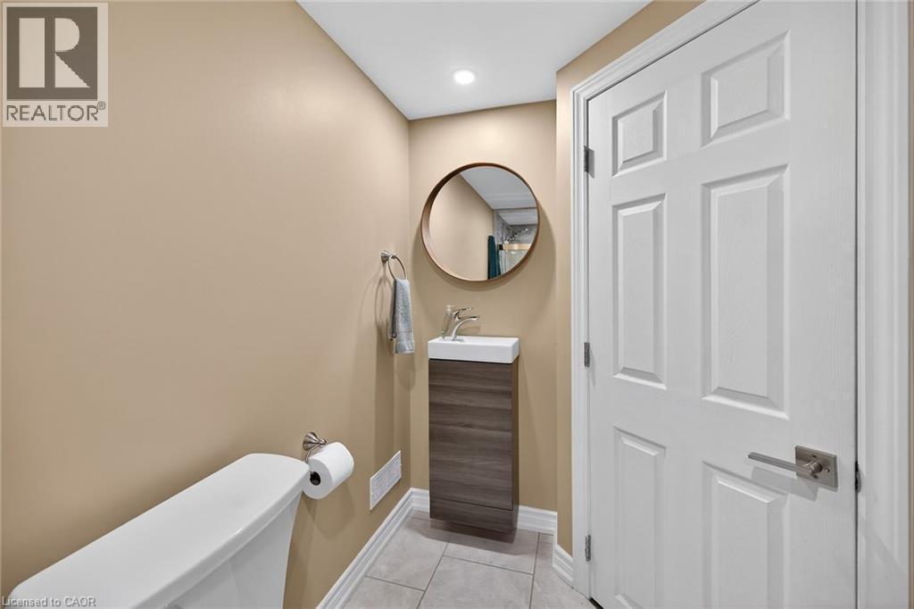 Contemporary powder room featuring a white toilet, pedestal sink with a rectangular basin, wood-look vanity, round mirror, and neutral wall tones - 149 Kinsman Drive, Binbrook, ON