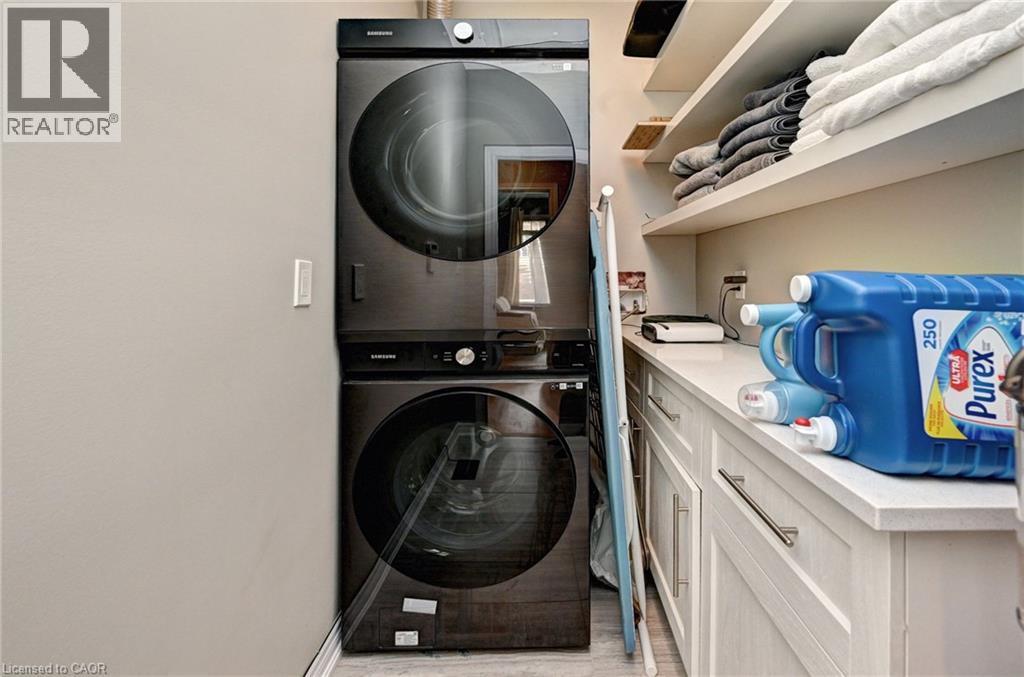 Stackable laundry appliances with a dark finish, white shaker-style cabinetry, light-toned countertop, and open shelving - 219 Falconridge Drive, Kitchener, ON - Indoor Photo Showing Laundry Room