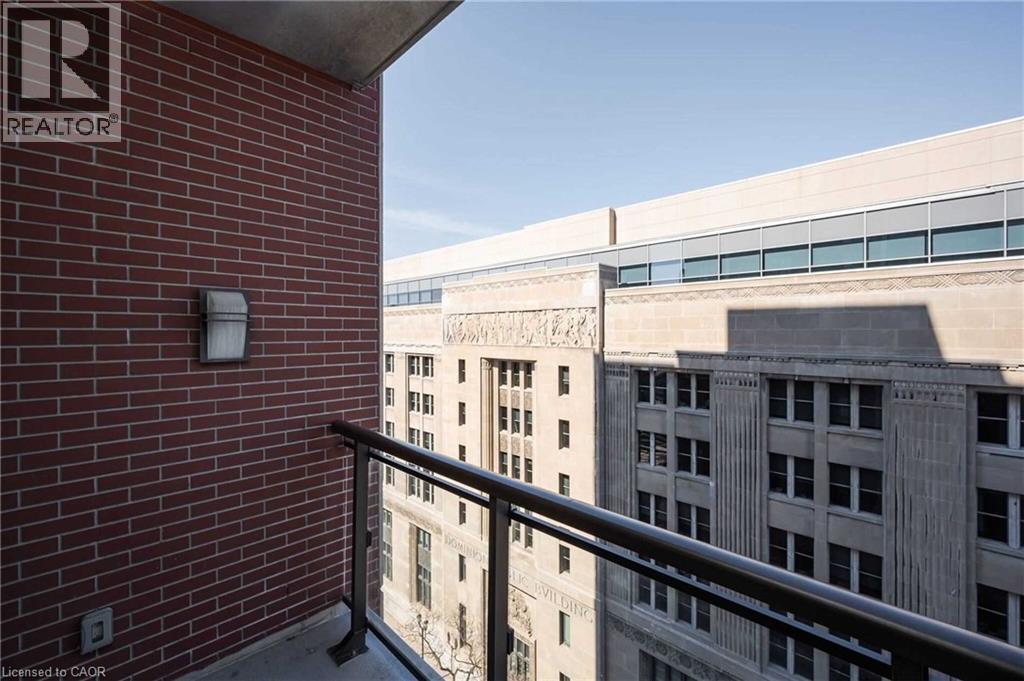Outdoor balcony featuring a metal railing, red brick wall, exterior light fixture, and concrete flooring - 112 King Street E Unit# 907, Hamilton, ON