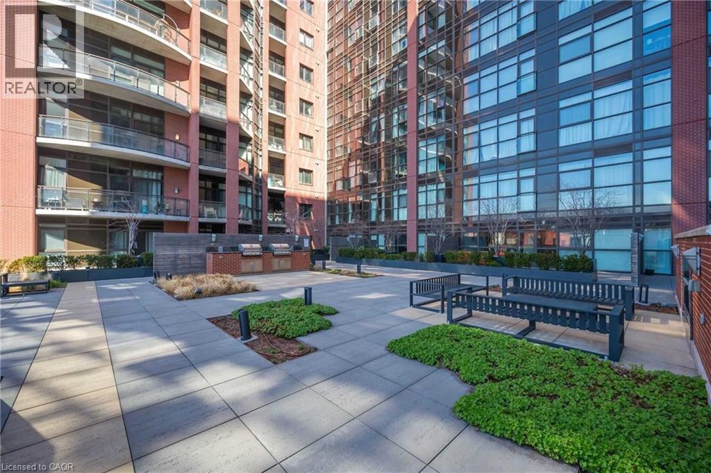 Spacious outdoor patio featuring paver flooring, integrated planters with greenery, multiple built-in grilling stations, and fixed bench seating - 112 King Street E Unit# 907, Hamilton, ON