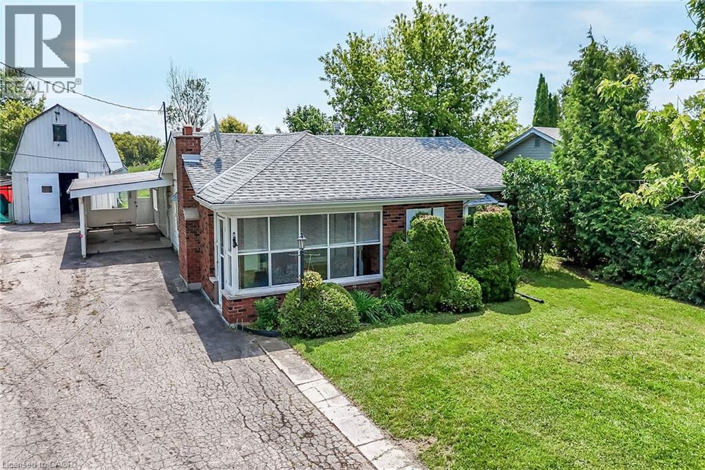 Brick-clad bungalow with a gray shingle roof, featuring a covered carport and a spacious front lawn with mature shrubbery - 114 E Second Road E, Stoney Creek, ON