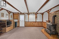 Room featuring a wood-burning stove with a brick surround, wood-paneled wainscoting, exposed wood beams, and a neutral carpet -