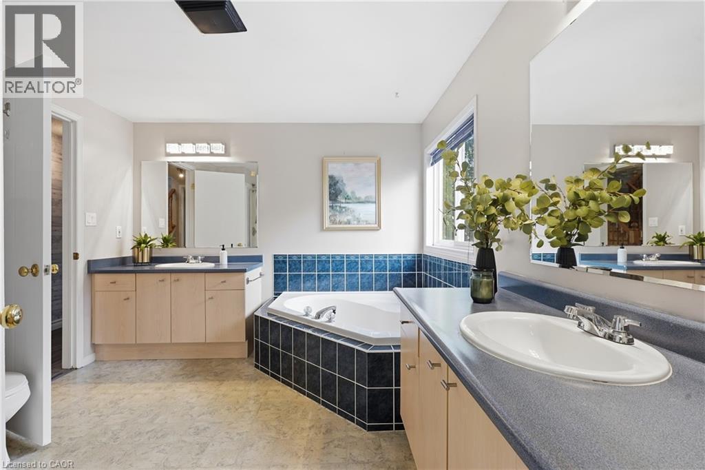 Bathroom featuring a corner soaking tub with dark tile surround, dual vanity with light wood cabinetry, gray countertops, and large wall-mounted mirrors - 128 Houghton Street, Cambridge, ON - Indoor Photo Showing Bathroom