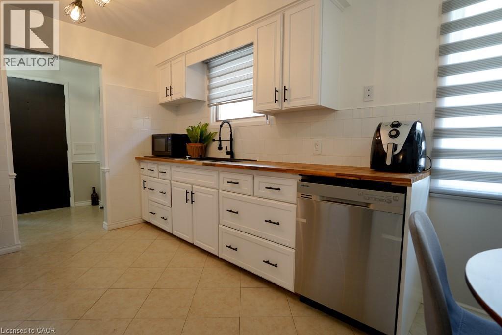 Kitchen featuring white cabinetry with matte black hardware, wood-finish countertops, stainless steel dishwasher, white subway tile backsplash, and a window with horizontal blinds - 147 Muriel Crescent, London, ON