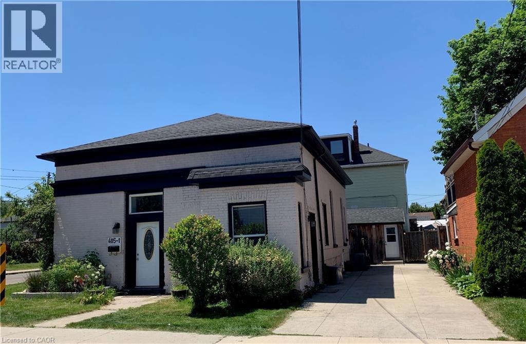 White brick exterior with a dark trim, featuring a white oval-lite entry door, a prominent front window, and a paved driveway - 405 Macnab Street N, Hamilton, ON