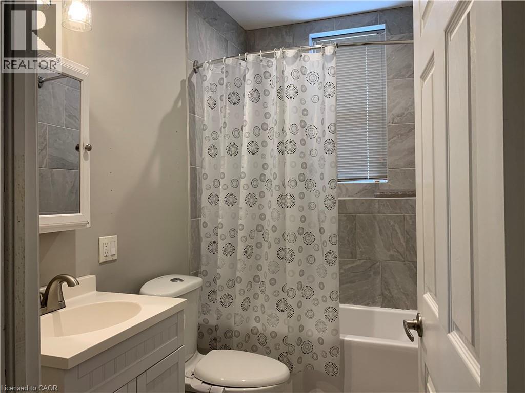 Unit 2 Bathroom featuring full tile surround, single vanity with integrated sink, panel cabinetry, medicine cabinet, and window with blinds - 405 Macnab Street N, Hamilton, ON