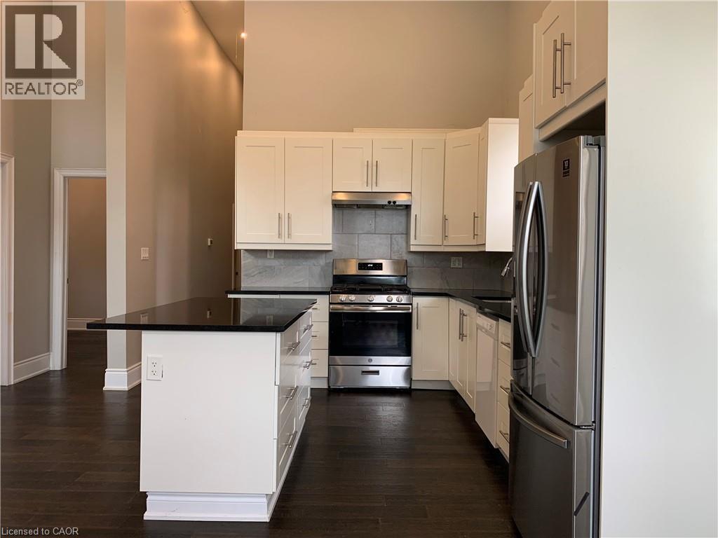 Unit 1Modern kitchen featuring an extended island with a black countertop, white shaker-style cabinetry, stainless steel appliances, and dark wood-finish flooring - 405 Macnab Street N, Hamilton, ON