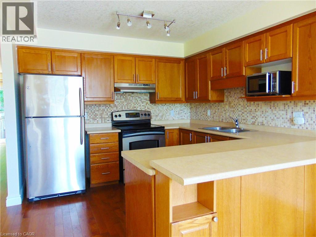 Kitchen featuring wood-finish cabinetry, stainless steel appliances, a mosaic tile backsplash, a double basin sink, and a breakfast bar peninsula - 214 Snowdrop Crescent, Kitchener, ON - Indoor Photo Showing Kitchen With Double Sink