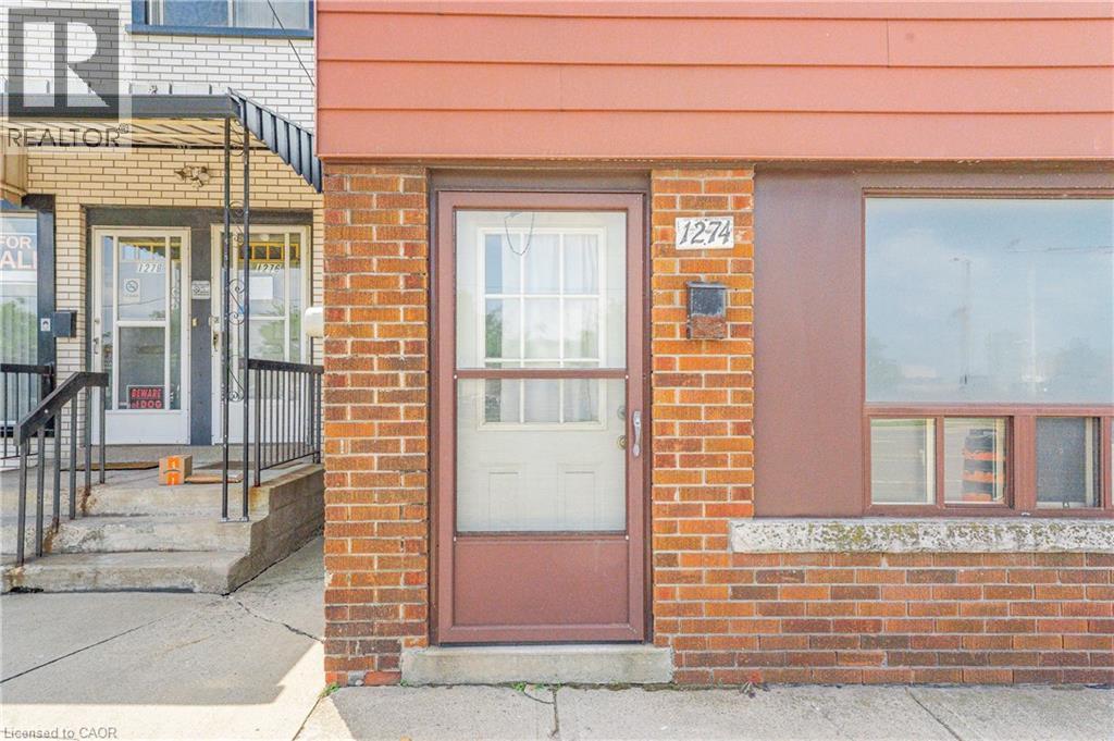 Brick facade building with a prominent front entrance featuring a storm door, an adjacent large pane window, house numbers, and a wall-mounted mailbox - 1274 Barton Street E, Hamilton, ON