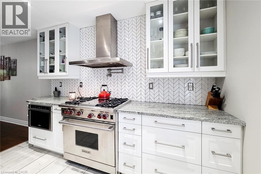 Kitchen featuring a stainless steel range with a wall-mounted range hood, granite countertops, white cabinetry with glass-front upper units, a herringbone tile backsplash, and wood-finish flooring - 29 Woodside Square, Fonthill, ON - Indoor Photo Showing Kitchen With Upgraded Kitchen